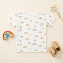 Neatly folded white baby T-shirt with pastel rainbow pattern, styled with dried chamomile flowers and a smooth pebble on a light wood background