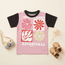 Neatly folded pink and black boys' T-shirt with floral graphic, styled with a small wooden toy camera, baby shoes, and dried flowers on a light neutral background