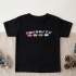 A flat-lay product photograph of a black toddler t-shirt with a colorful pastel 'FAVORITE' text and icon graphic, presented on a beige linen surface with miniature holiday trees and pinecones.